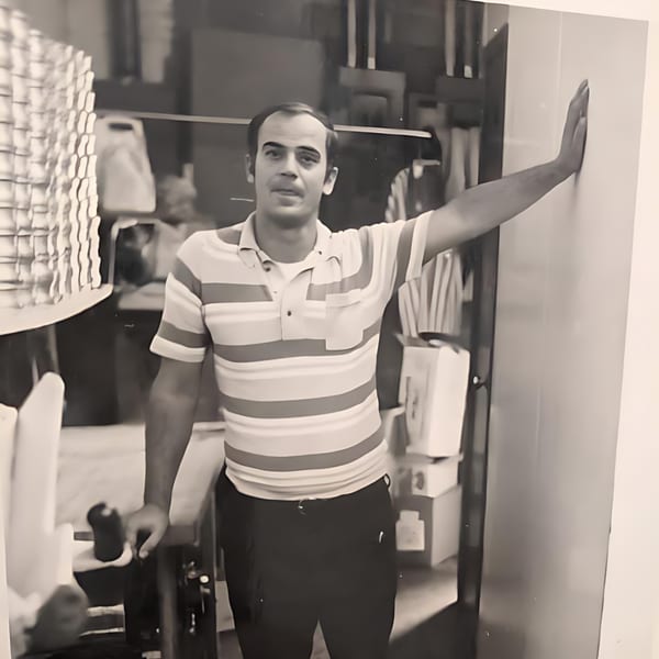 Black and white photo of a man in a striped shirt leaning against a wall in a kitchen.