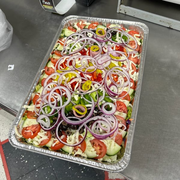 Large catering tray of fresh salad with lettuce, tomatoes, cucumbers, onions, olives, and cheese.