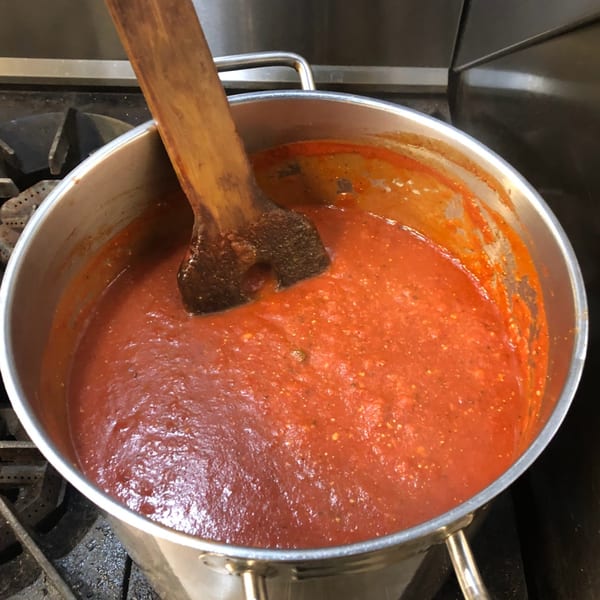 Large pot of simmering red pizza sauce with a wooden spoon on a stove.