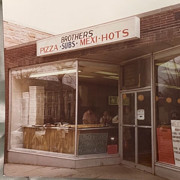 Exterior of Original Brothers Pizza, a brick building with a sign advertising pizza, subs, and Mexi-Hots.